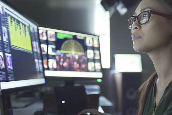 Close up stock image of a young asian woman sitting down at her desk where she‚Äôs surrounded by 3 large computer monitors displaying out of focus images of people as thumbnails; crowds; graphs & scrolling text.
